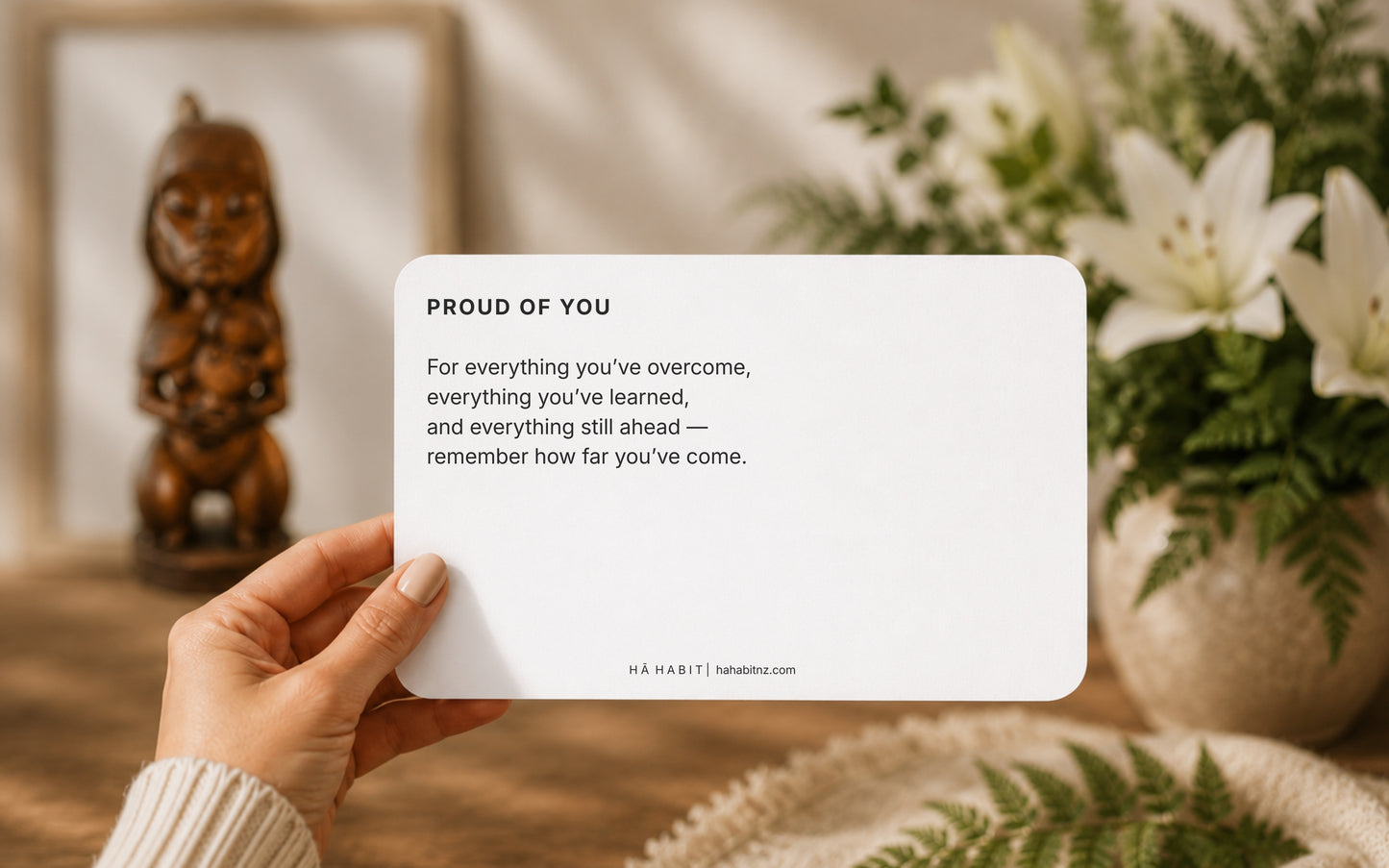 A hand holds a white rectangular card with the text 'PROUD OF YOU' and a supportive message, set against a warm, softly blurred background with a wooden figurine and white lilies. Proud of you" card — for everything you've overcome and everything still ahead