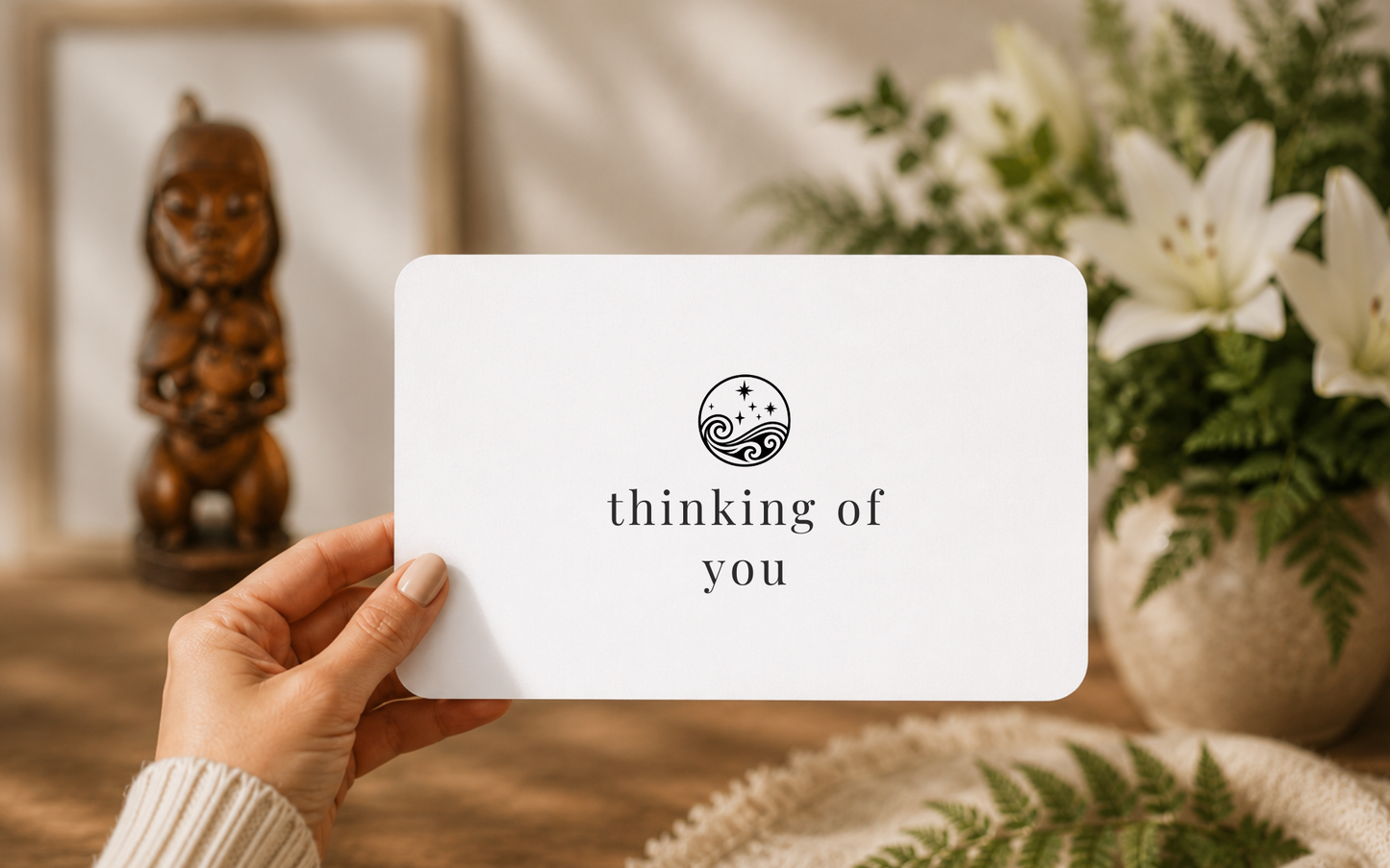 A hand holds a white rectangular card with a black circular moana/sea and stars/whetu logo and the text 'thinking of you' in a warm indoor setting with flowers and a wooden figurine in the background.