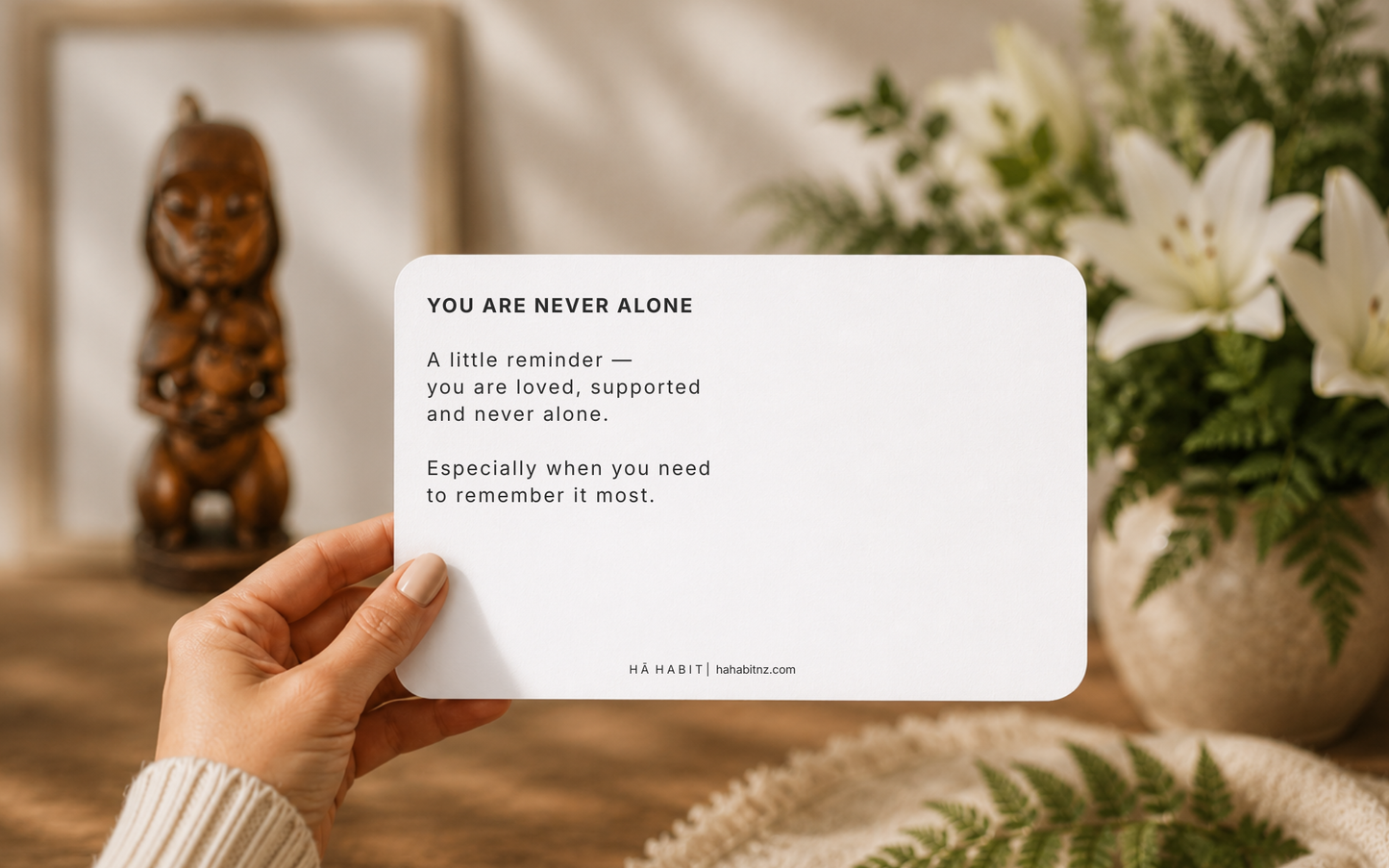 A hand holds a white rectangular card with printed positive affirmation text reading 'YOU ARE NEVER ALONE' and smaller supporting copy. The background is a softly blurred warm indoor scene with flowers, greenery, and a wooden figurine.